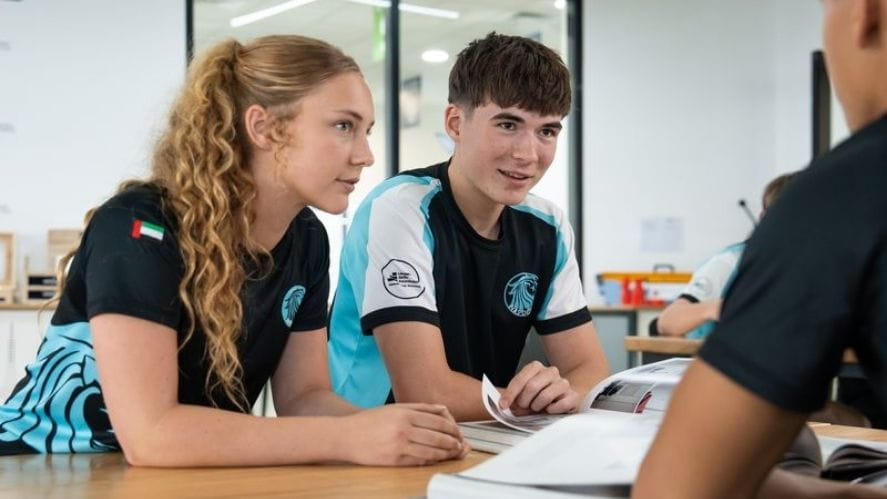 Engaged Student Learning Two NAS Dubai students in school sports uniforms sitting at a classroom table, engaged in discussion and working from open books.