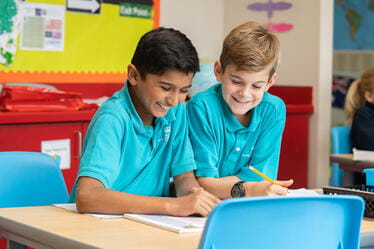 Two students seated at a table, writing and smiling