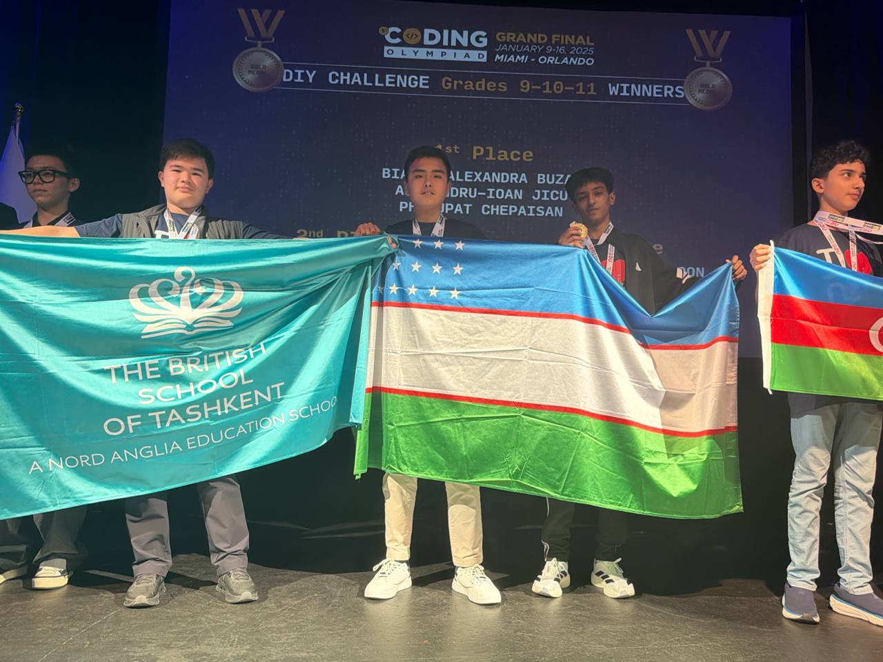 A group of BST participants stand on a stage holding large flags including The British School of Tashkent flag and the Uzbekistan national flag.