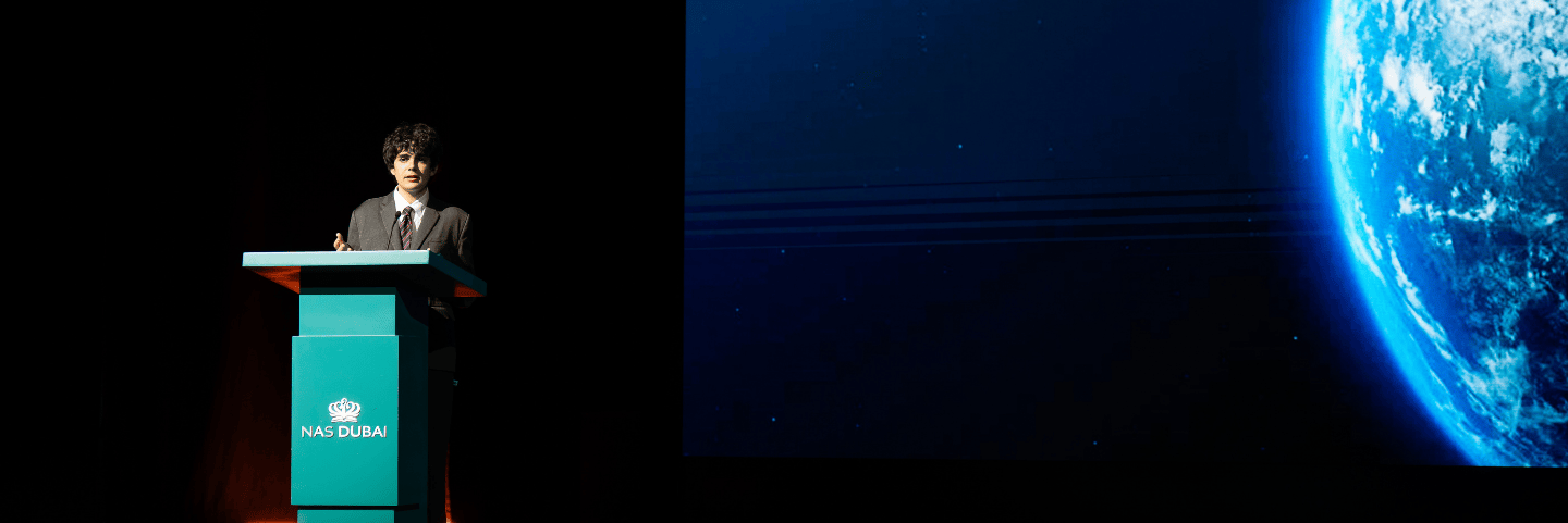 Sixth Form standing at an NAS Dubai podium on a dimly lit stage with a large projected image of Earth visible on the right side of the screen.