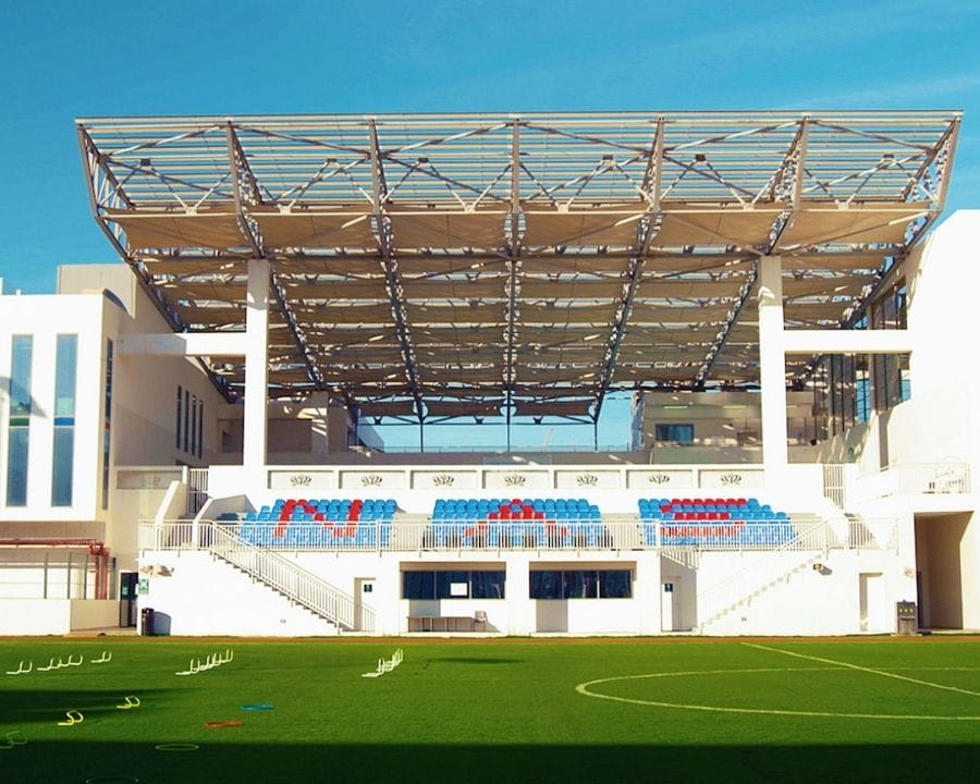 Full-sized sports pitch at NAS Dubai with a modern covered grandstand, red and blue seating, surrounding white school buildings, and training equipment on the grass under a clear blue sky.