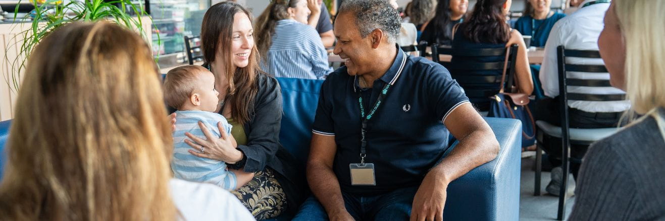 Group of parents seated together in a bright café-style setting at NAS Dubai, holding a baby and engaging in conversation during a community meetup, with other families in the background.