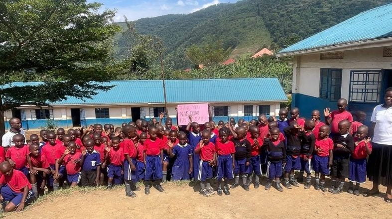 A large group of children in matching red and navy uniforms stand outdoors in front of blue‑roofed school buildings. The children hold a pink sign that reads 'Thank You NAS Dubai,' with green hills and trees visible in the background.