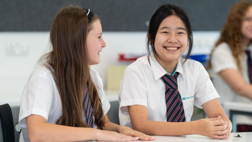 Students in uniform seated at a table in a NAS Dubai classroom during a lesson, with additional students in the background.