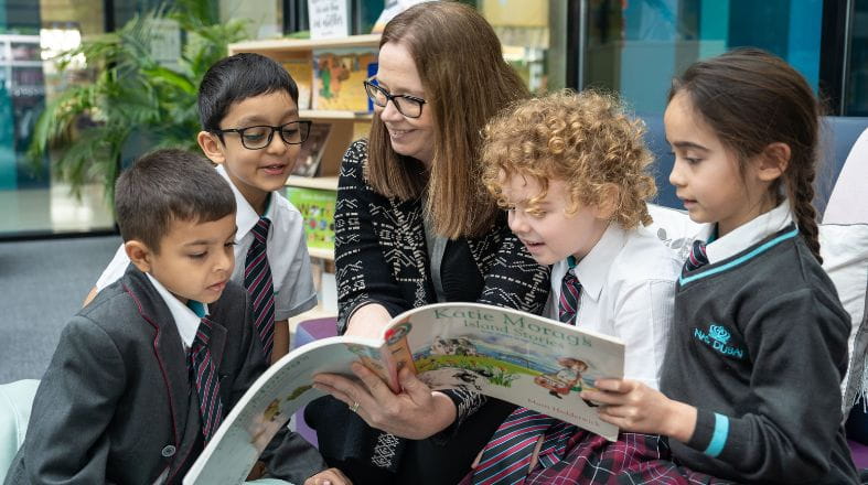 A teacher sits with a group of NAS Dubai students in the school library, guiding them through a large picture book. The children gather closely around, following along as bookshelves, plants, and soft seating create a welcoming reading environment.