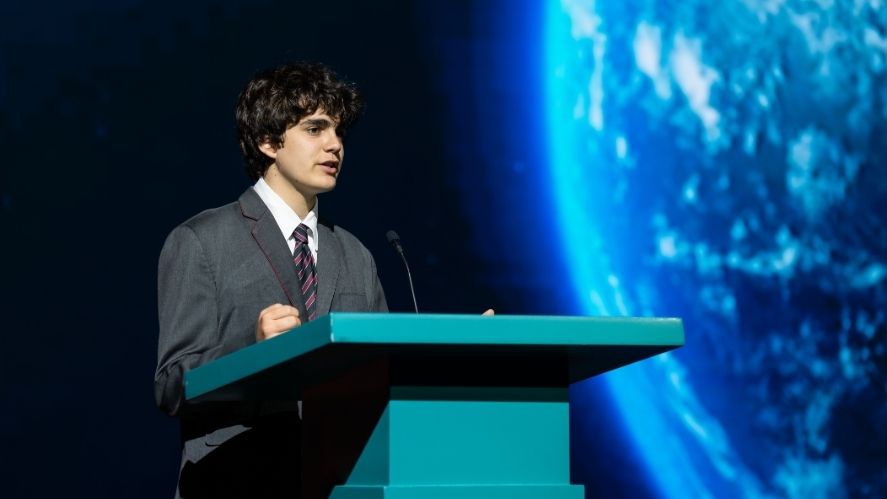 NAS Dubai student confidently speaking on stage at a school event, standing behind a NAS Dubai podium.