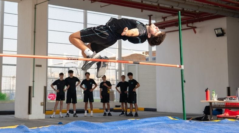 Student performing a high jump while classmates watch and cheer from the side.