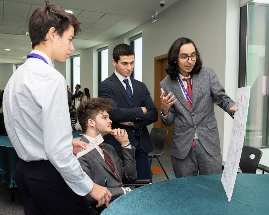Sixth form students gathered around a table as one student presents information from a display board during an academic presentation session.
