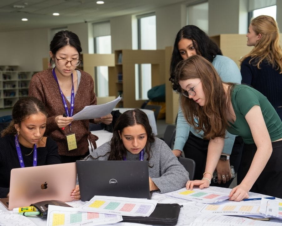 Sixth form students gathered around a table with laptops and printed documents, collaborating on a group project in a bright study area.