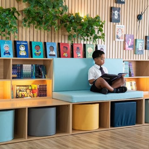 Student reading a book in a bright school library, seated on a cushioned bench with bookshelves, colorful book covers, and soft storage cubes in the background.