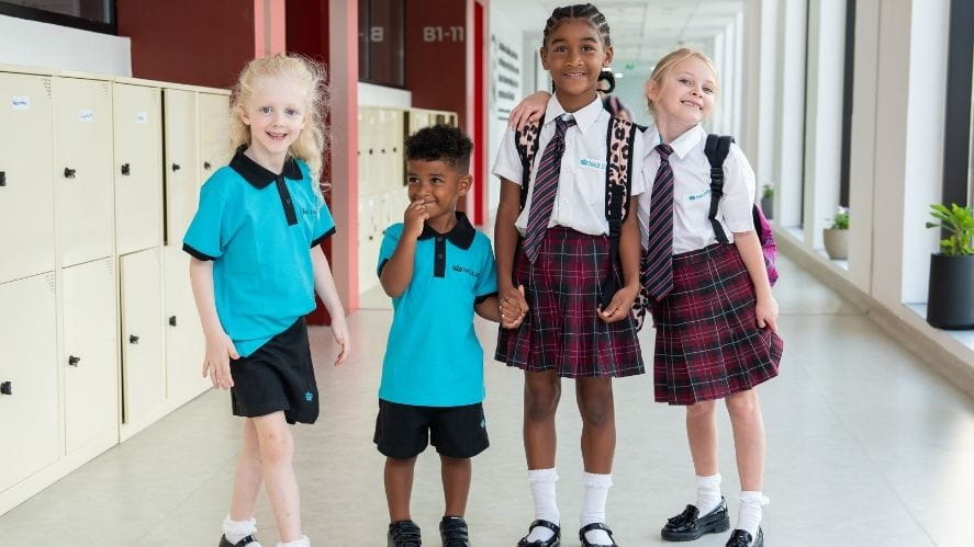 A group of NAS Dubai students A group of four NAS Dubai students stand together in a bright school corridor lined with lockers. Some students wear turquoise PE uniforms while others wear the school’s plaid uniform with white shirts and ties. They appear to be interacting and holding hands as they prepare for their school day.