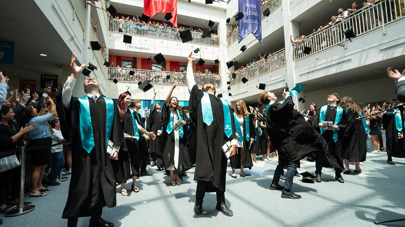 Graduates in black gowns and teal stoles throw their caps into the air inside a spacious atrium, surrounded by cheering spectators on multiple balconies.
