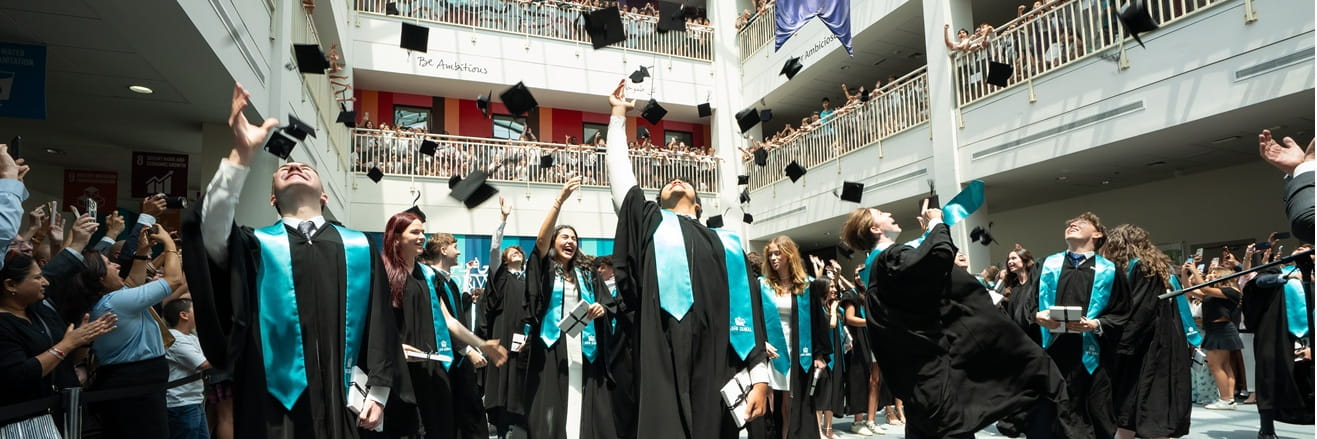 Graduates in black gowns and teal stoles throw their caps into the air inside a spacious atrium, surrounded by cheering spectators on multiple balconies.