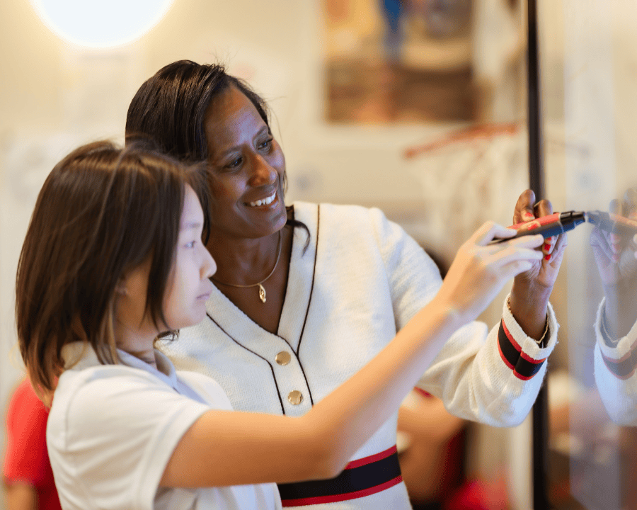 Middle school math teacher smiling and guiding a student as they write on a smartboard in a classroom setting.