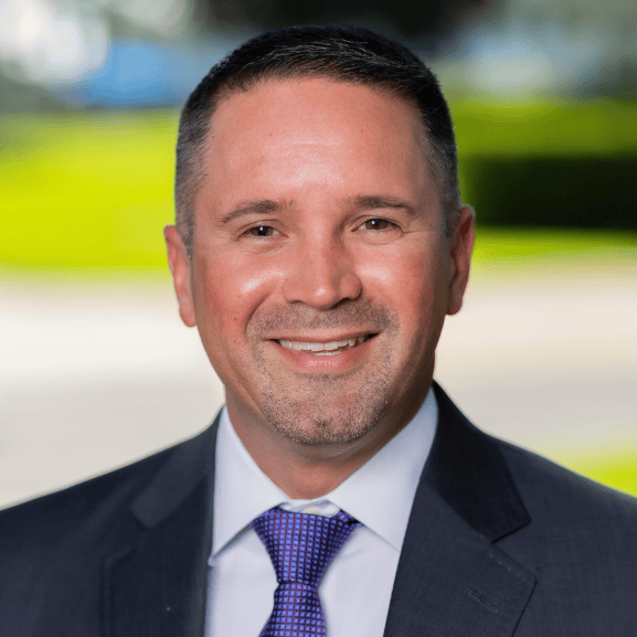 Professional headshot of a WPS Interim Head of School in a dark suit posed in front of a softly blurred outdoor background with greenery