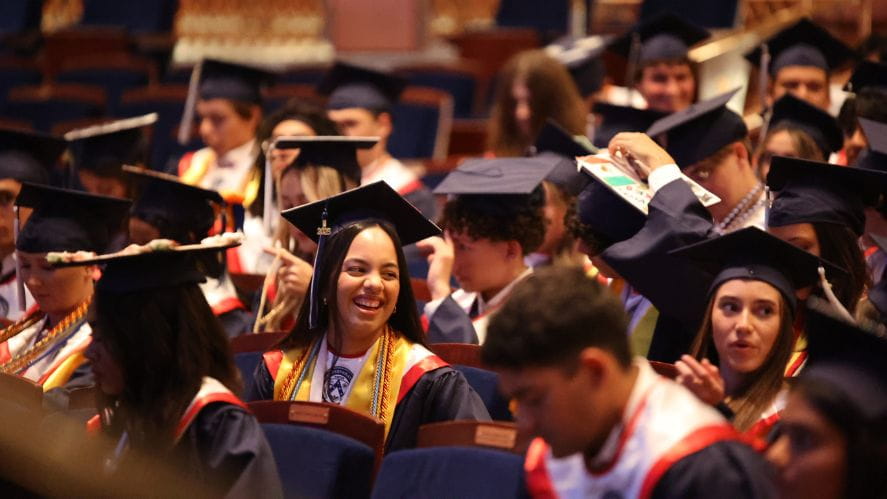 High school graduates in caps and gowns seated in an auditorium, smiling and chatting as they prepare for the ceremony.