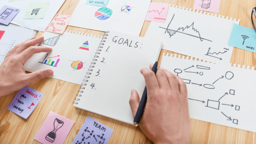 Student writing goals in a notebook surrounded by charts, graphs, and planning notes on a desk.